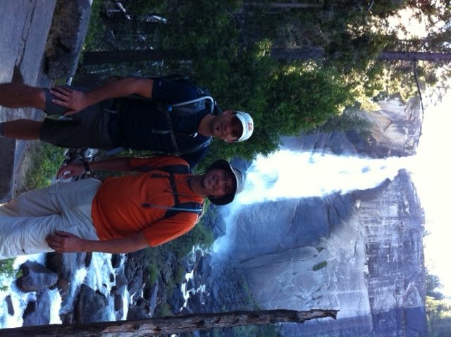 Two men pose near a waterfall; one in orange shirt, the other in a blue shirt, smiling in nature.
