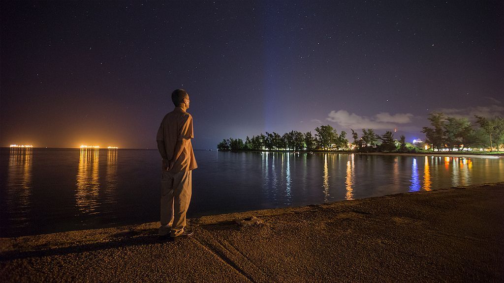 Person gazing at starry night sky over water, with lights of buildings reflecting.