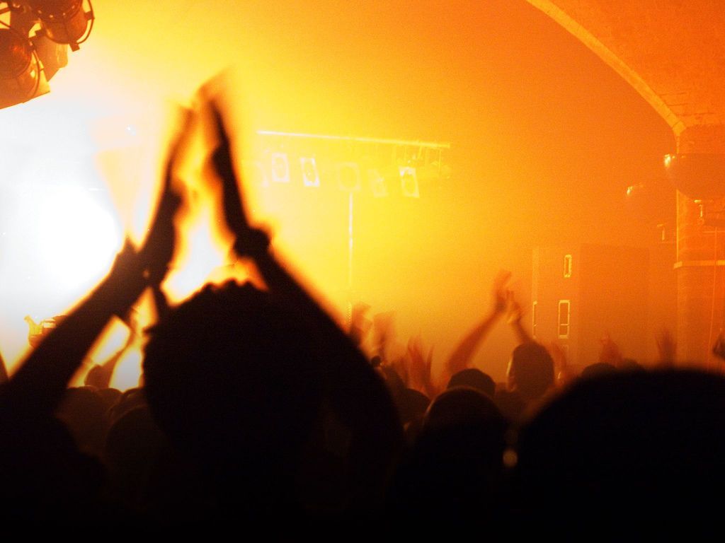 People clapping at a concert lit by bright orange stage lights.