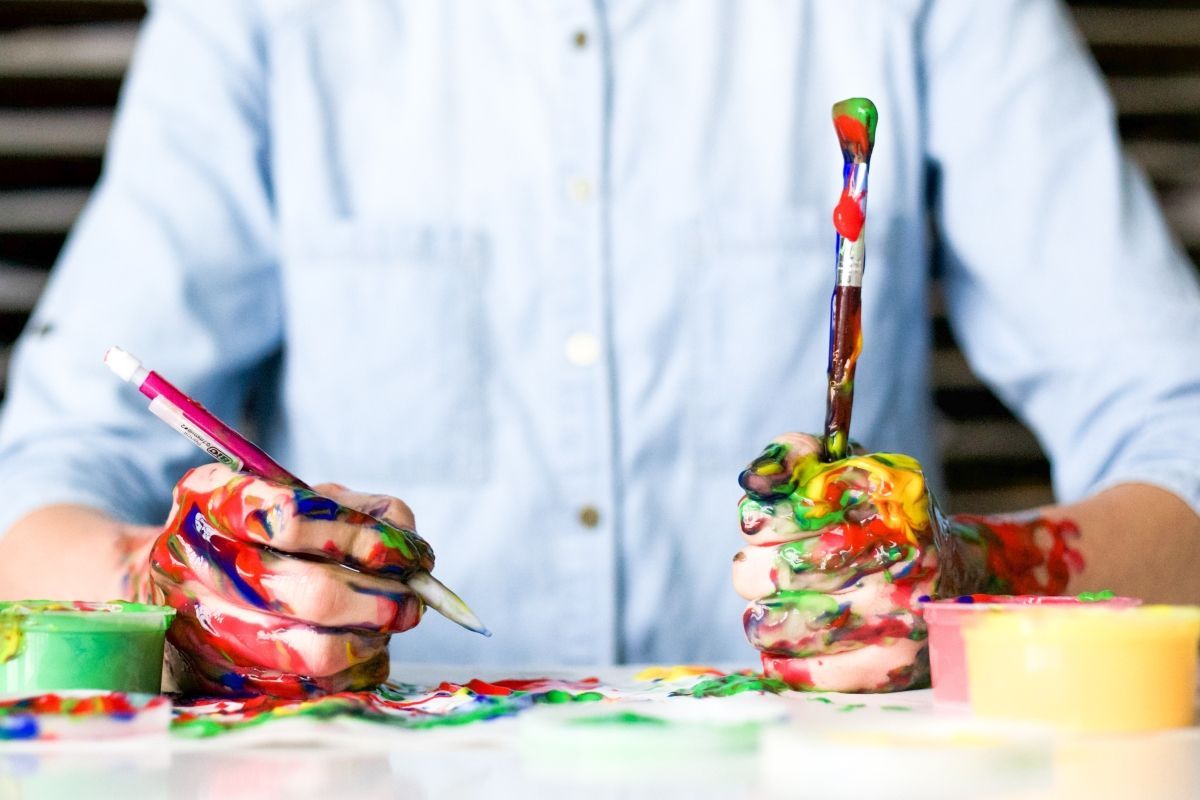 Hands covered in paint holding a paintbrush and pen, working on a project.