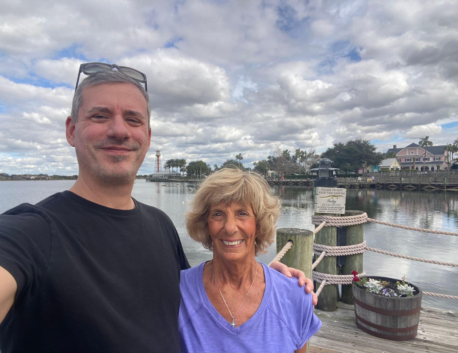 Two people smiling on a waterfront dock with boats, cloudy sky, and harbor buildings in the background
