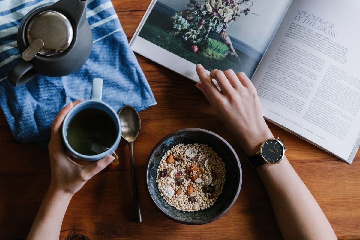 Person's hands holding a mug of tea and reading a magazine over a bowl of cereal with a watch on their wrist.