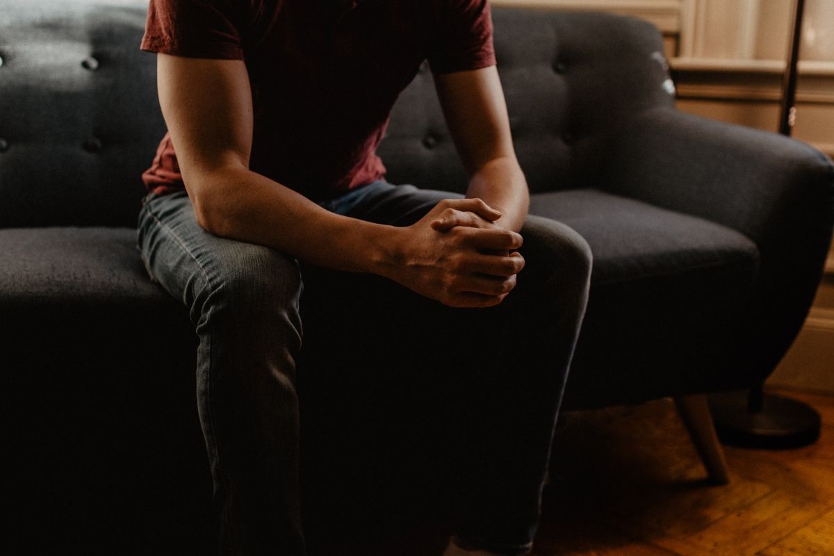 Man sitting on a dark gray couch with clasped hands. He is wearing a maroon shirt and jeans.