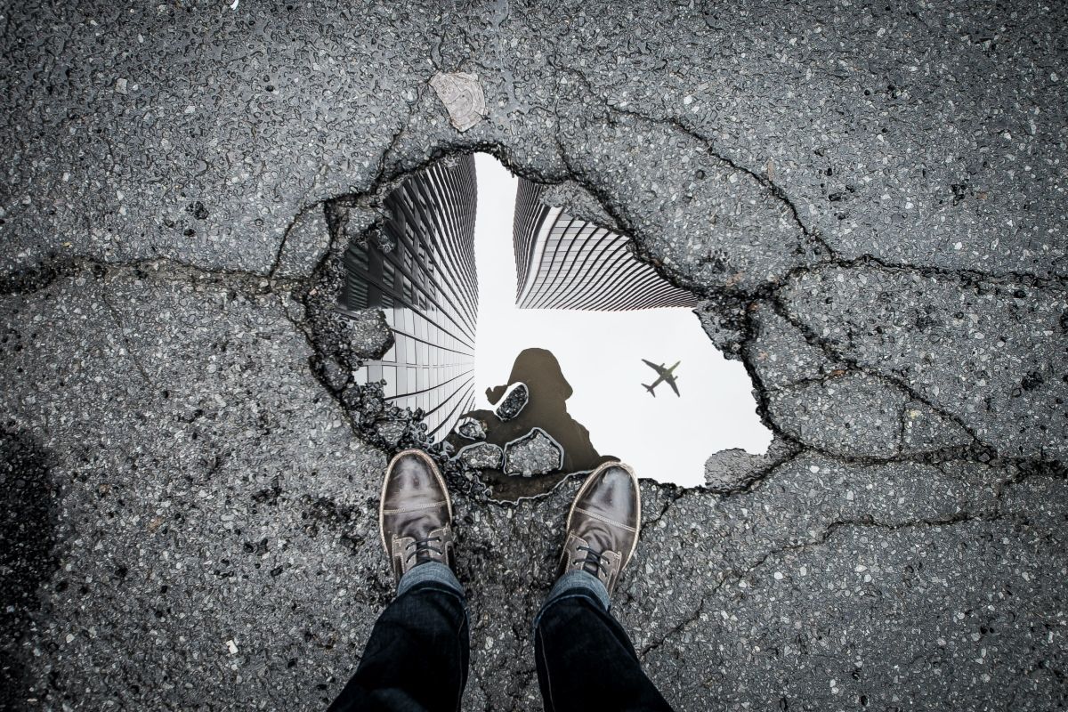 Person standing over a puddle reflecting a building and a plane in asphalt.