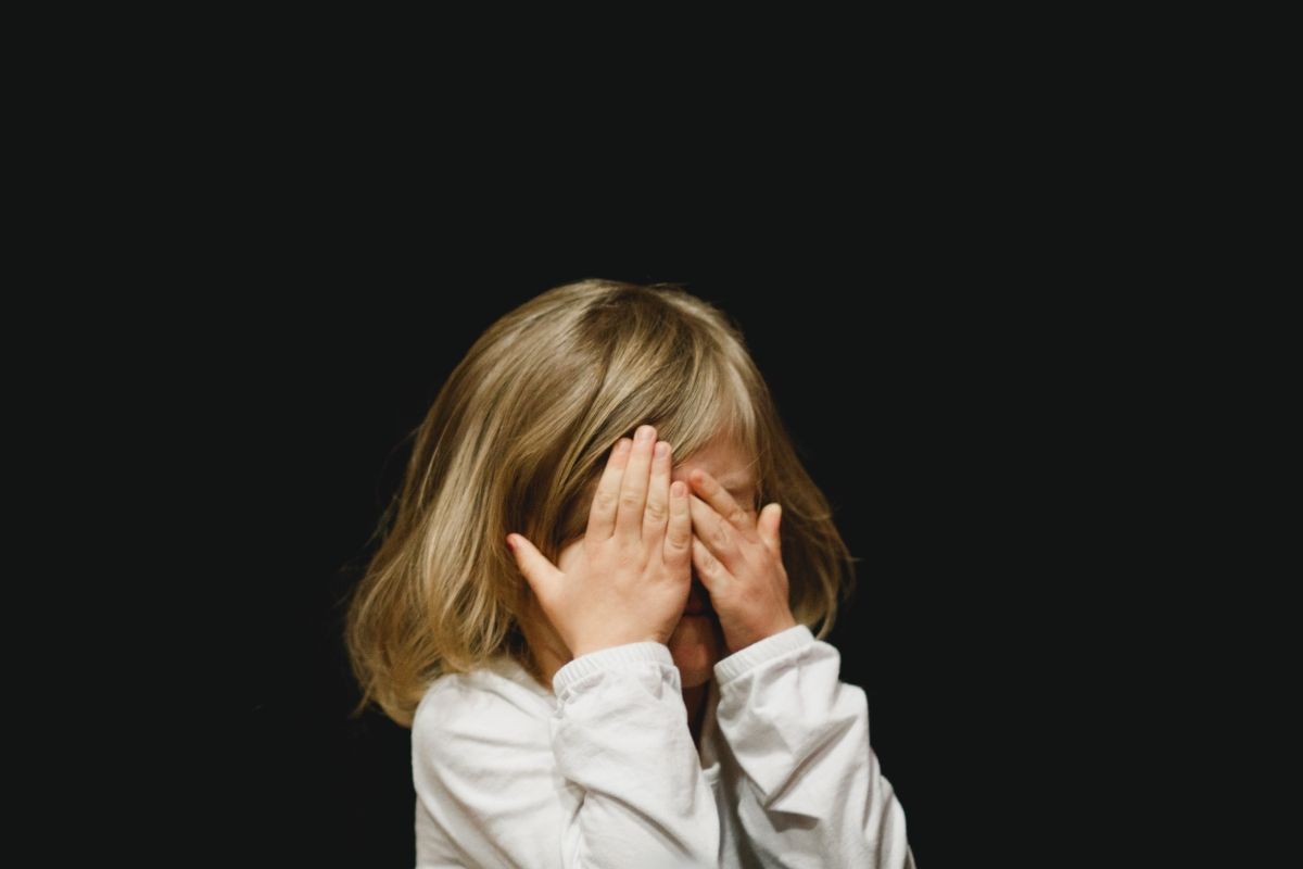 Blonde child covers eyes with hands, standing against a black background.