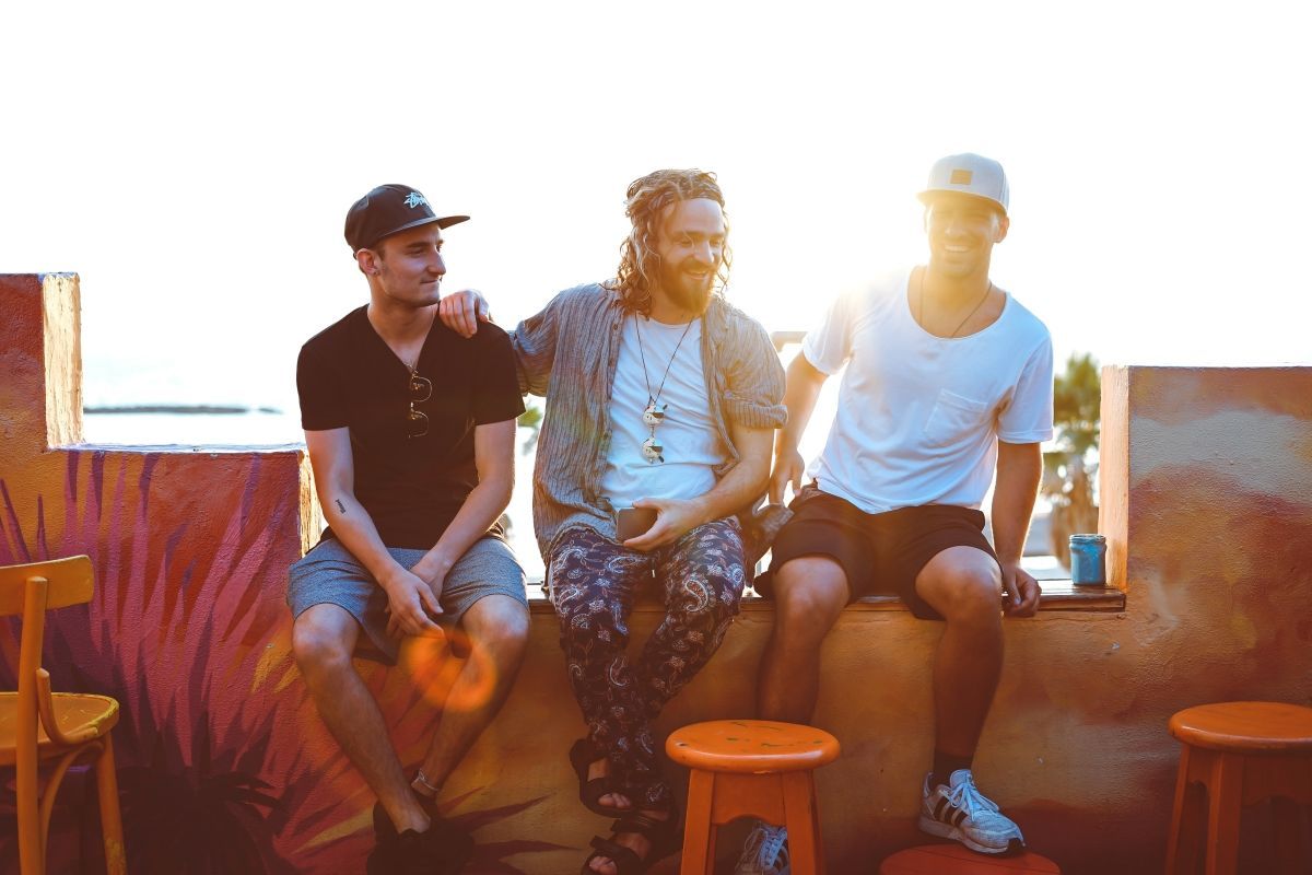 Three men sit outdoors on a ledge, smiling in the sun. One has dreadlocks, the others wear caps.