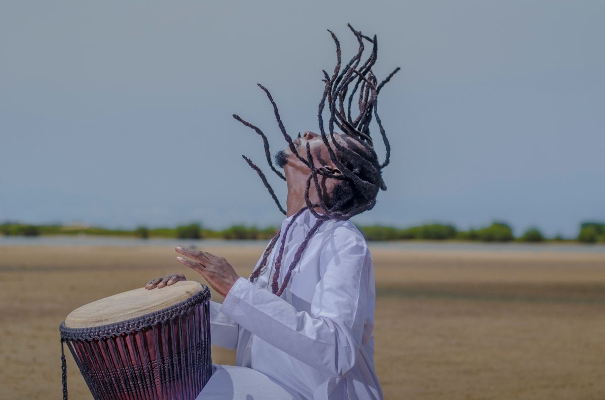 Person playing a drum with dreadlocks flying up, outdoors. Wearing white robe. Dry, sandy landscape and sky in background.