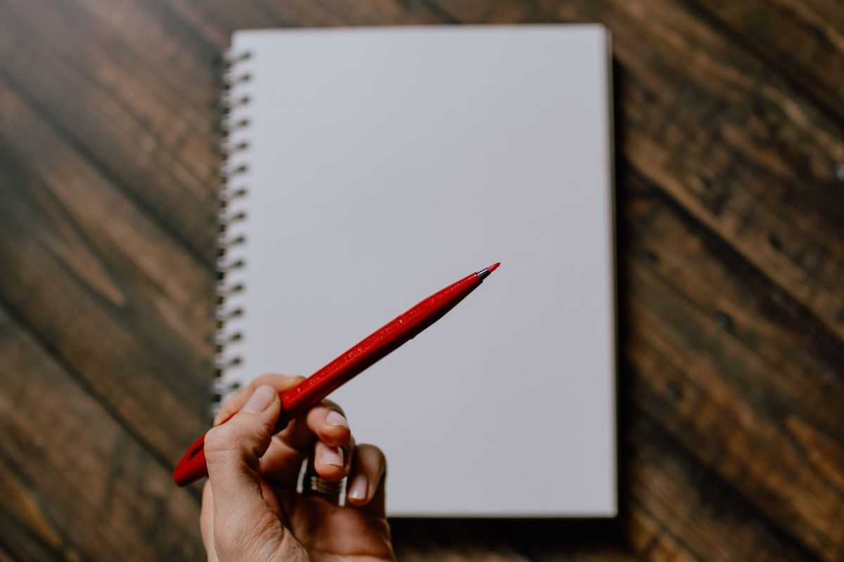 Hand holding a red pen over an open, blank spiral notebook on a wooden surface.