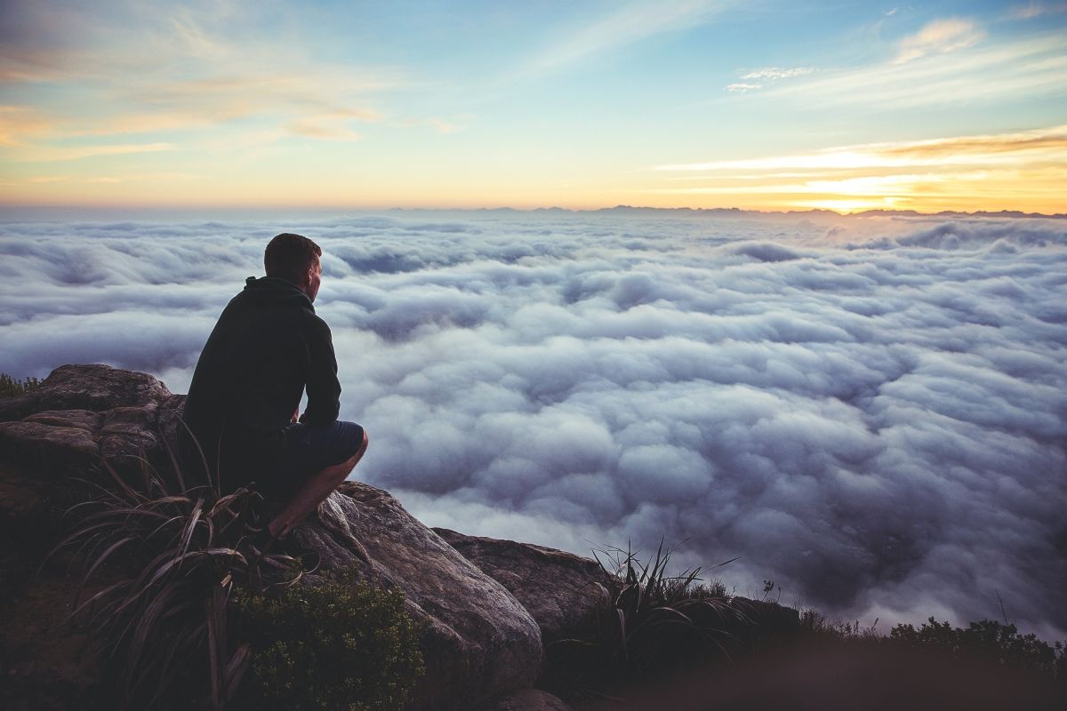 Man sits on cliff, gazing over a sea of clouds at sunrise, orange and blue sky.