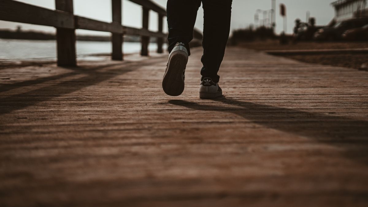 Person walking on a wooden pier towards the horizon; sun casts long shadows, dark clothing.