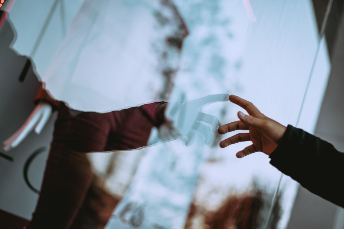 Hand reaching towards a person reflected in glass, holding a sign. The person wears dark clothing.