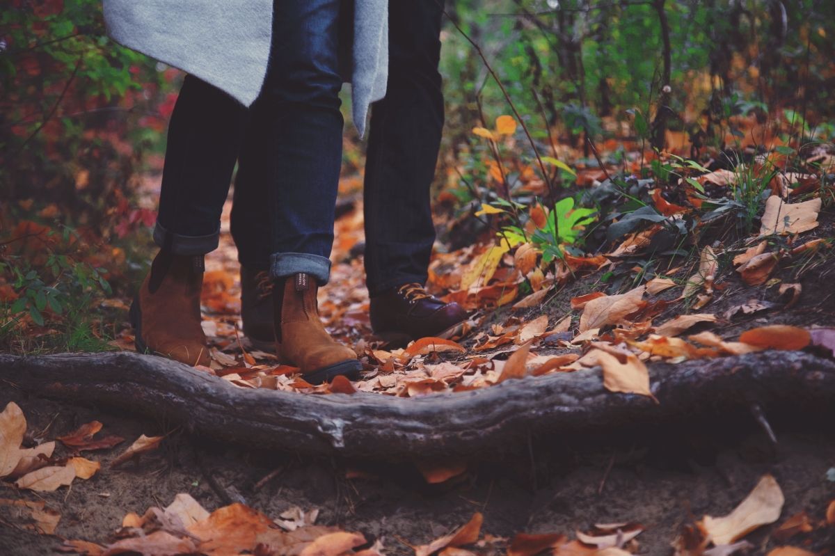 Couple standing close together on a trail covered in autumn leaves; brown boots and jeans visible.
