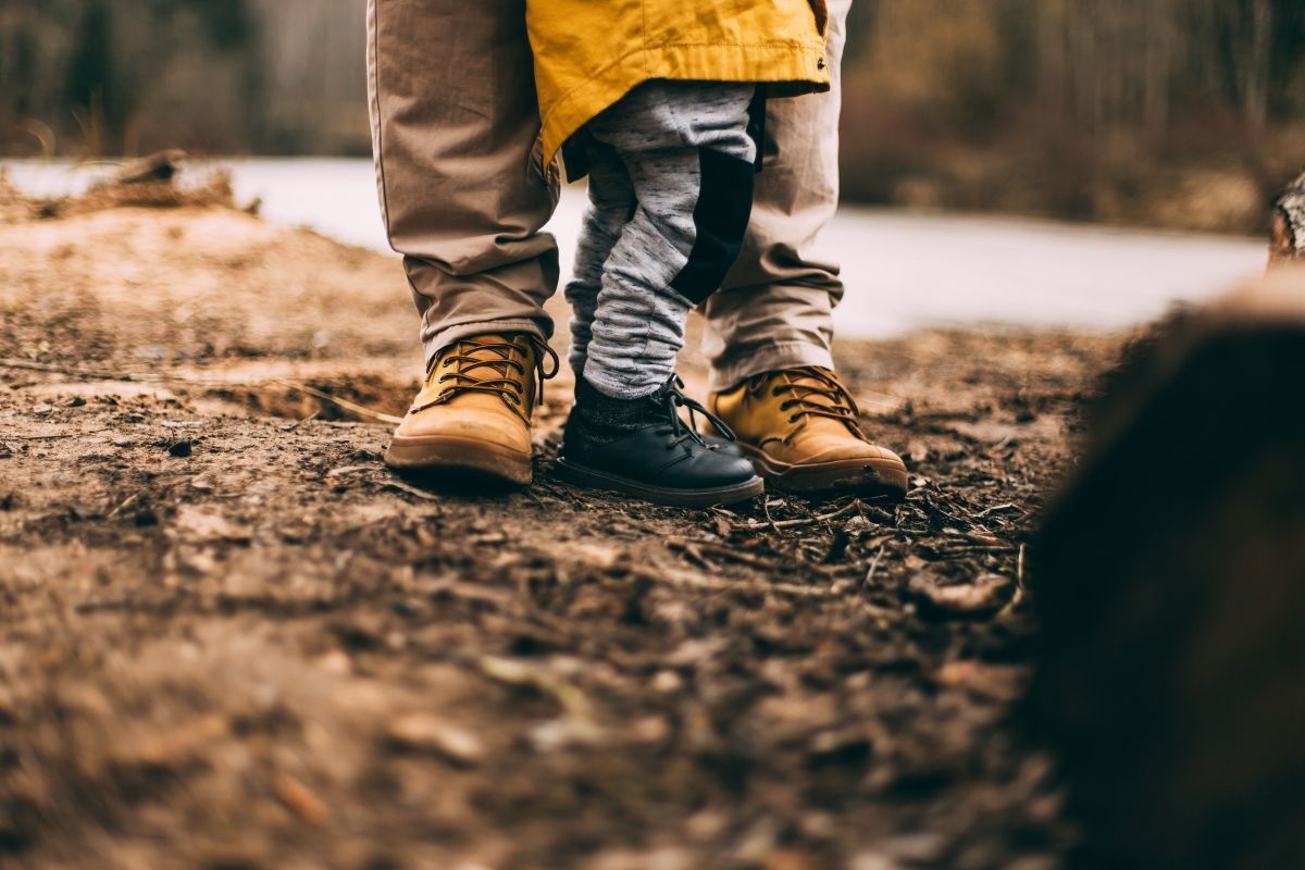 Adult and child's boots stand side-by-side on a brown, earthy ground. The adult is wearing tan boots. The child has blue.
