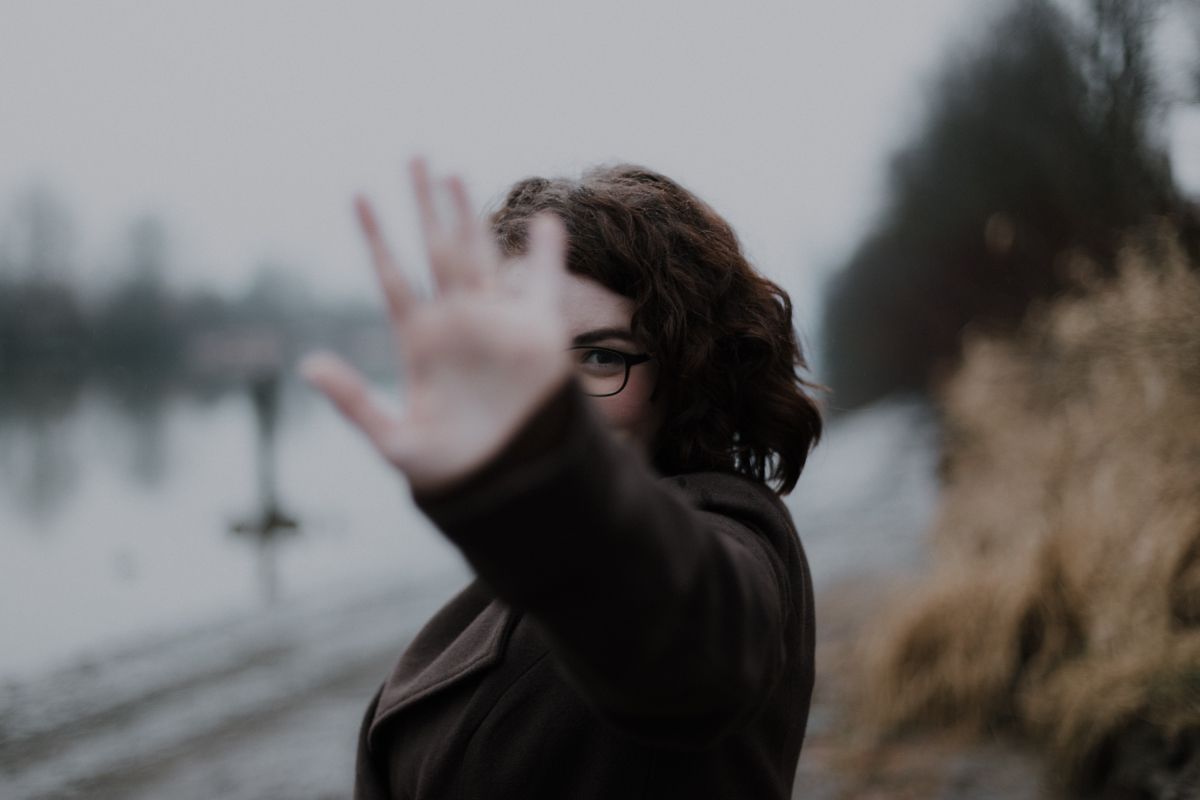 Woman with glasses holds up hand in front of face, blurring the view; outdoors, brown coat.