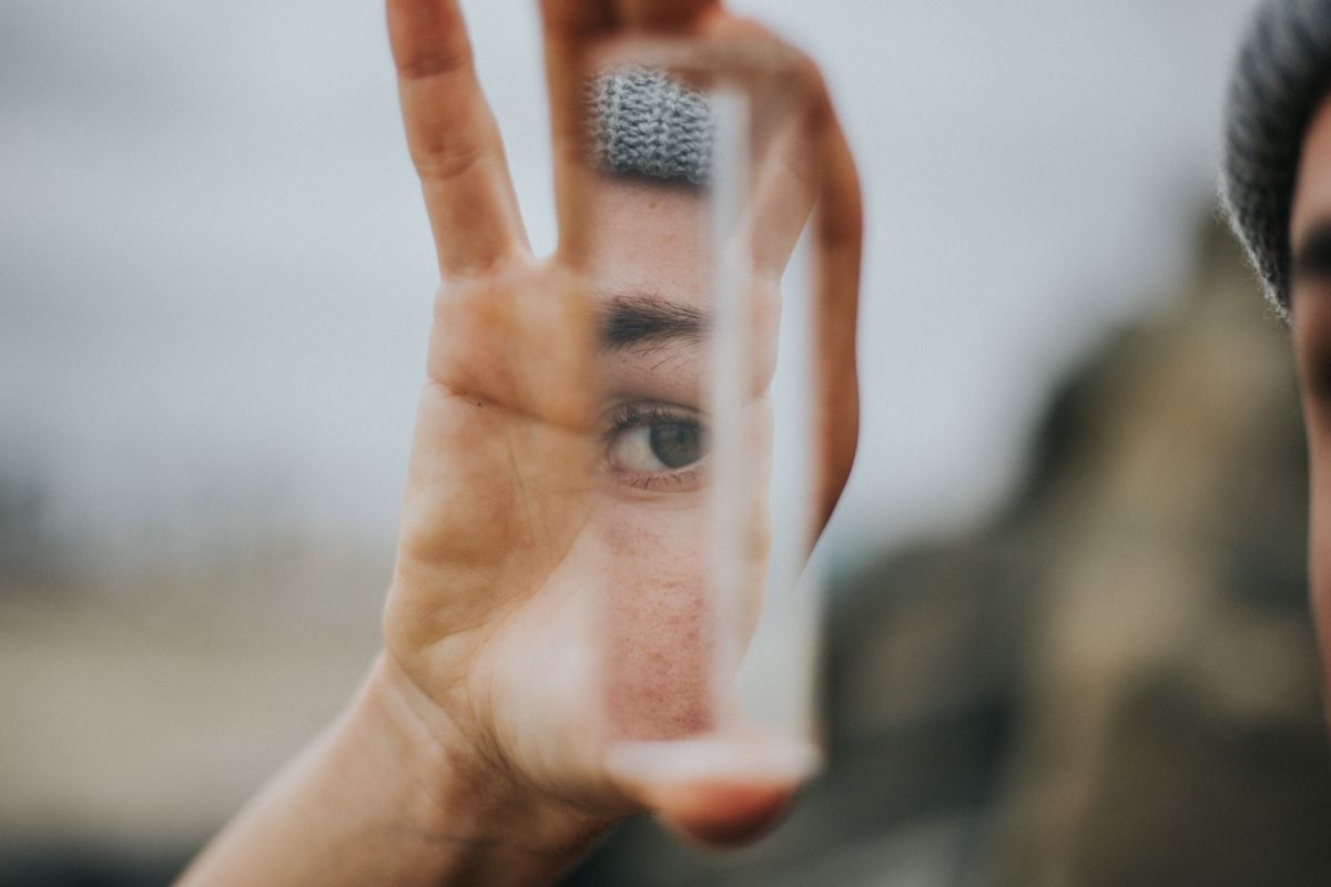 Hand holding a small mirror, reflecting a close-up of an eye against a blurry outdoor background.