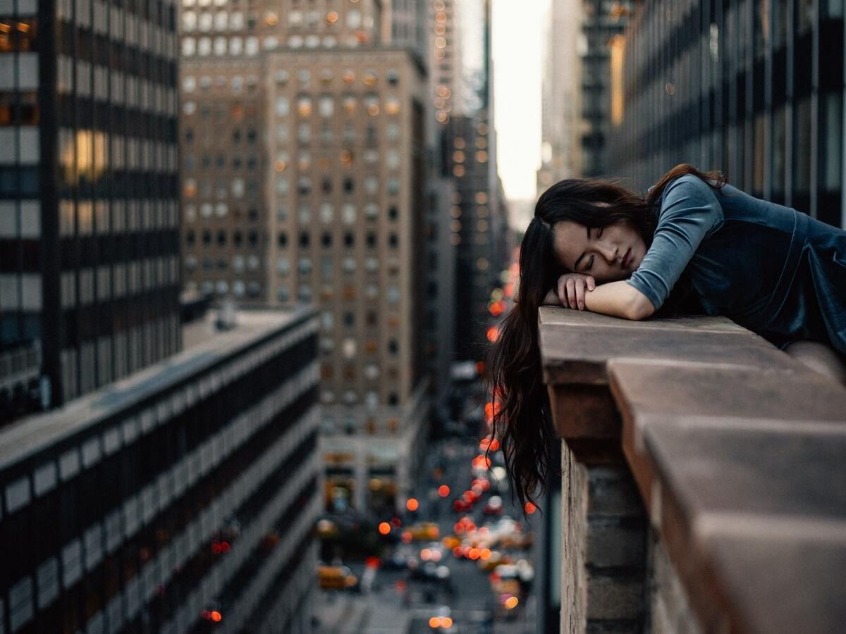 Woman resting head on rooftop ledge, city view below.
