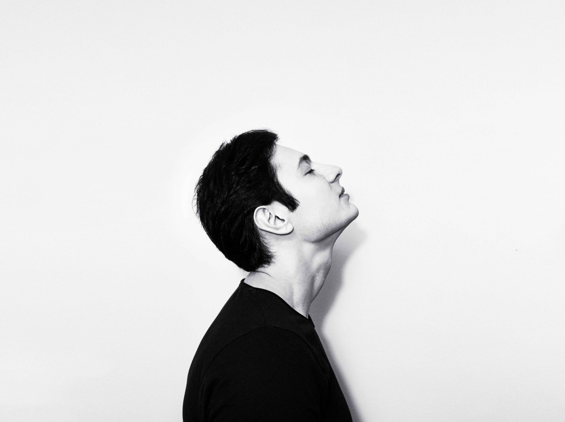 Man with dark hair looks upwards, eyes closed, in black shirt against a white background.