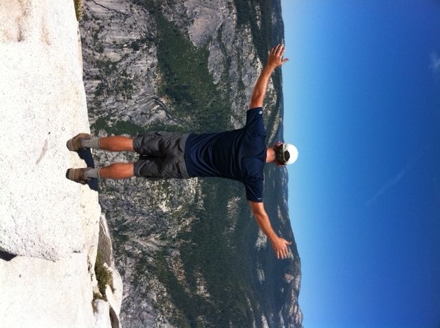 Man on cliff edge with arms outstretched, blue sky background, green valley below.