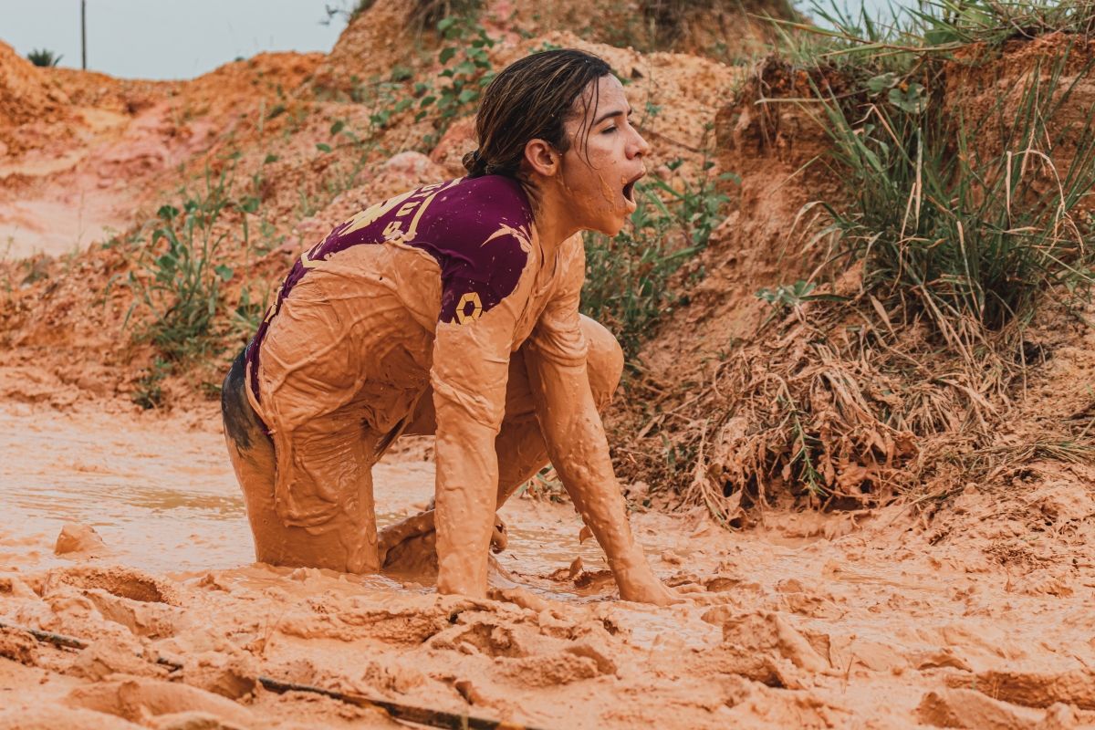 Woman covered in mud, screaming, in a reddish-brown muddy area, with vegetation nearby.