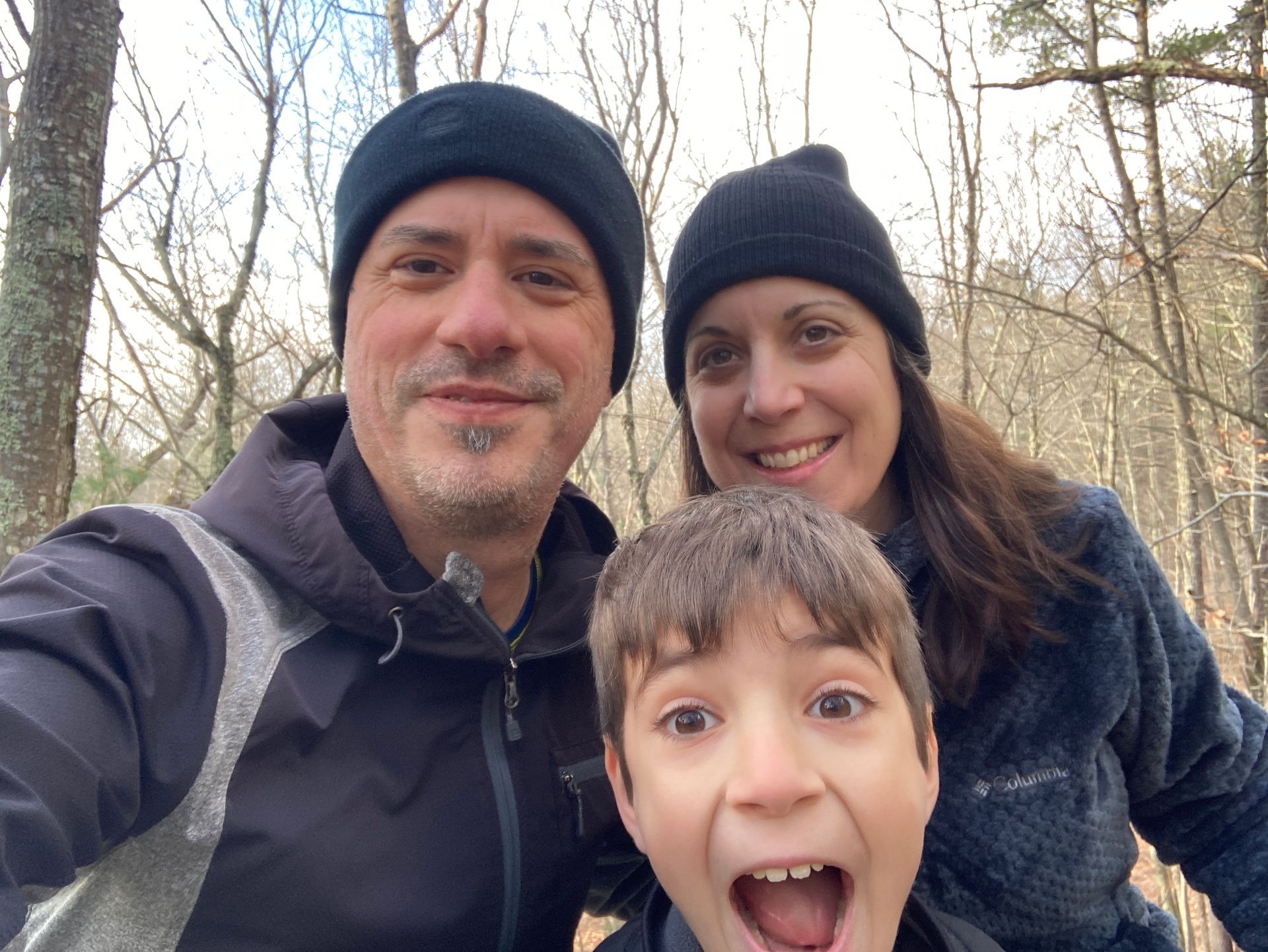 Three people smiling outdoors in winter hats and jackets, with a child in front making a playful face