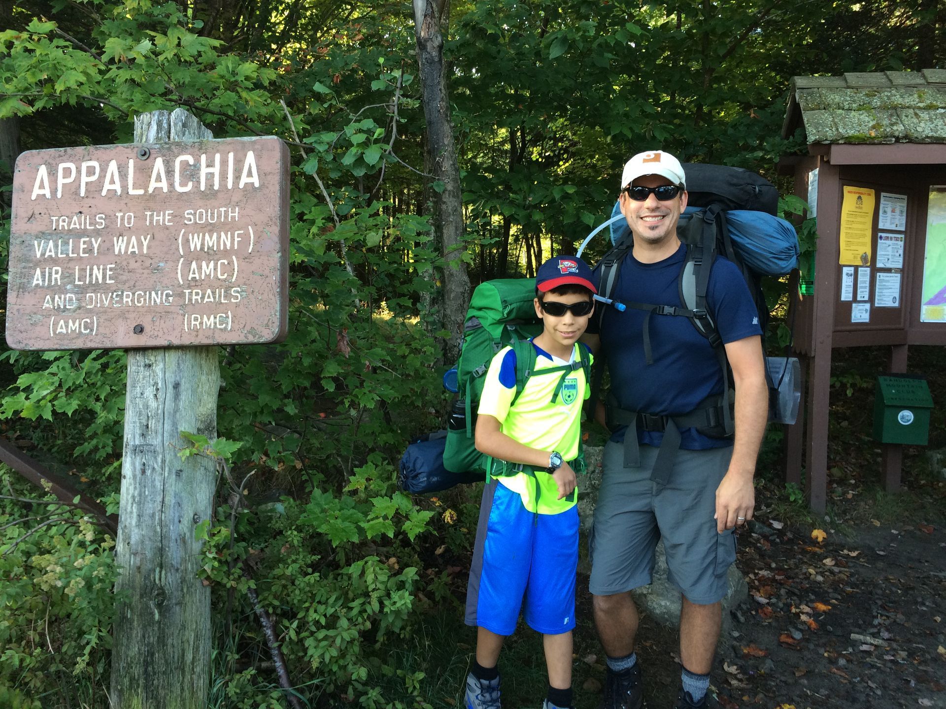 Two hikers with backpacks posing beside an Appalachia trail sign in a wooded area