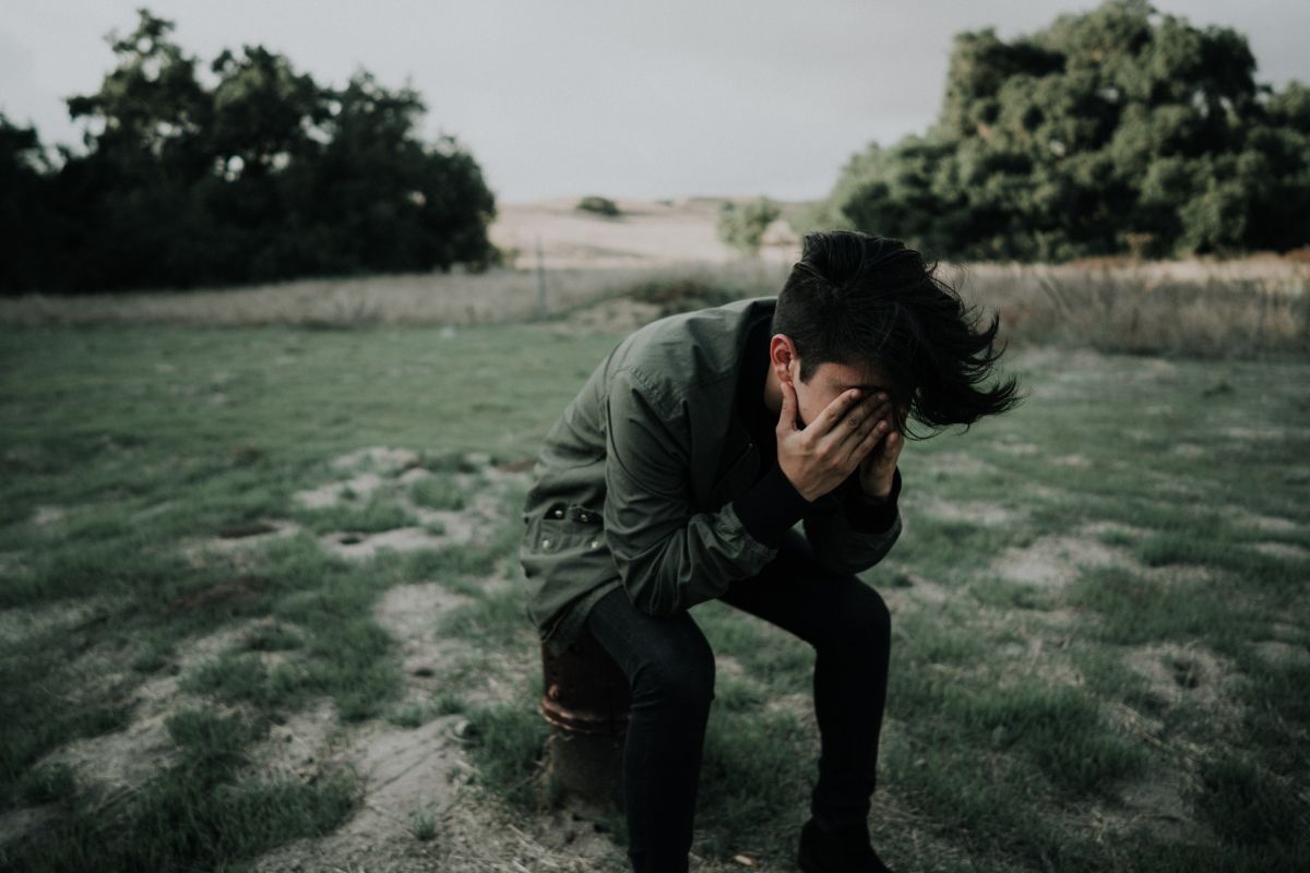Person sitting outside, covering face with hands, appearing sad or upset. Overcast sky, green grass, trees.