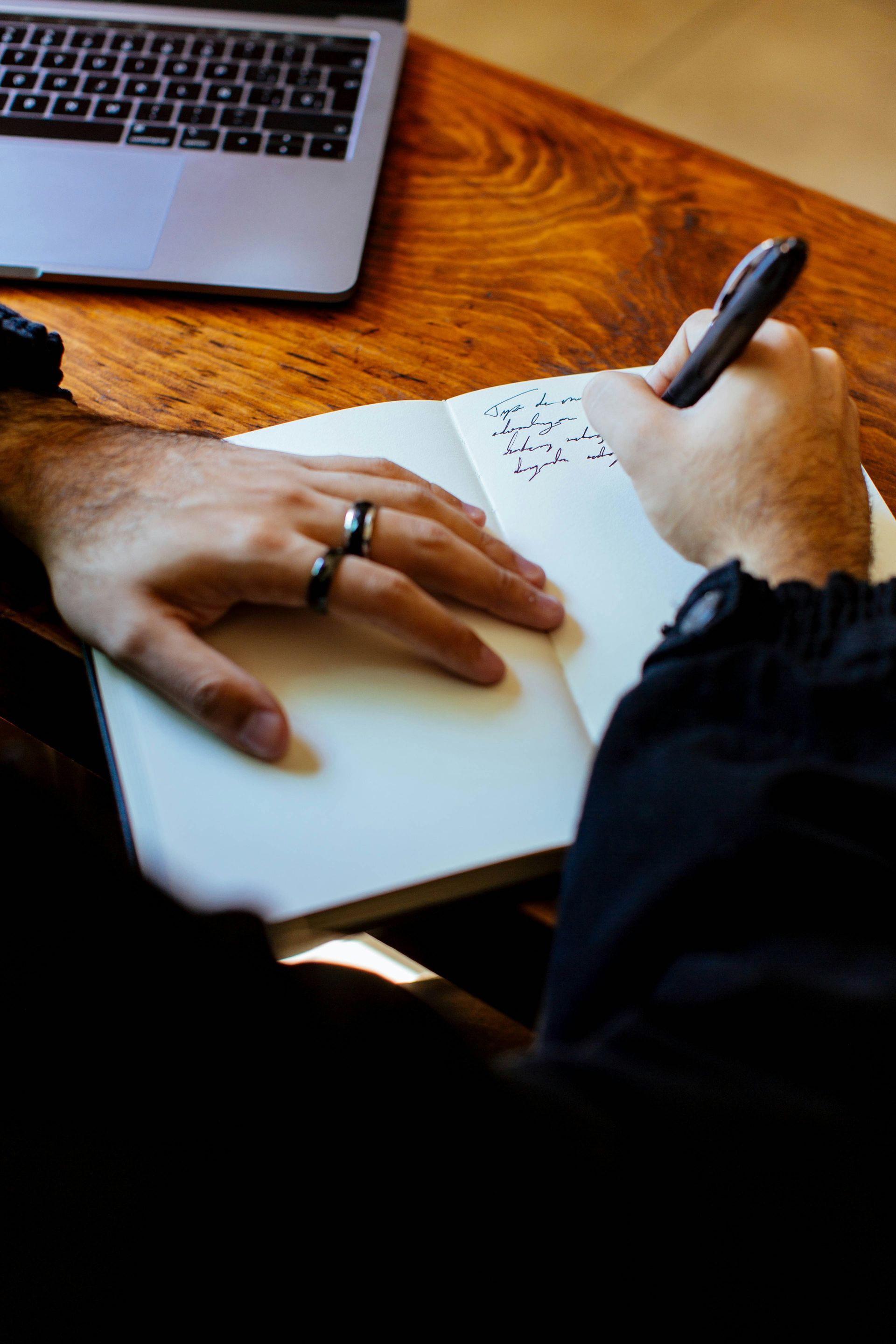 Hands writing in a notebook beside a laptop on a wooden table