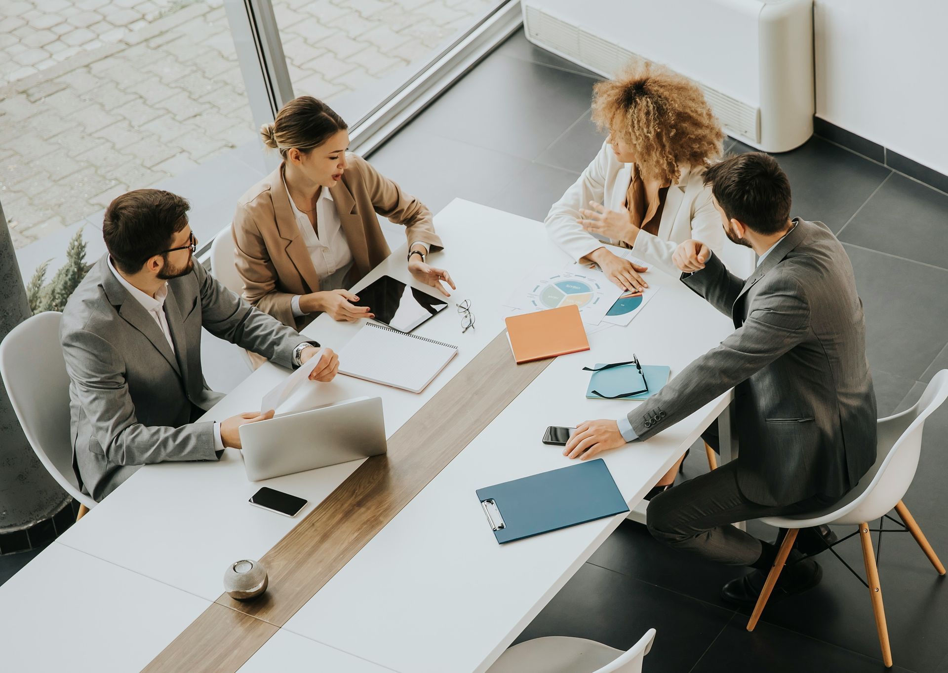 Four coworkers in a bright office meeting around a white table with papers, notebooks, and laptops.