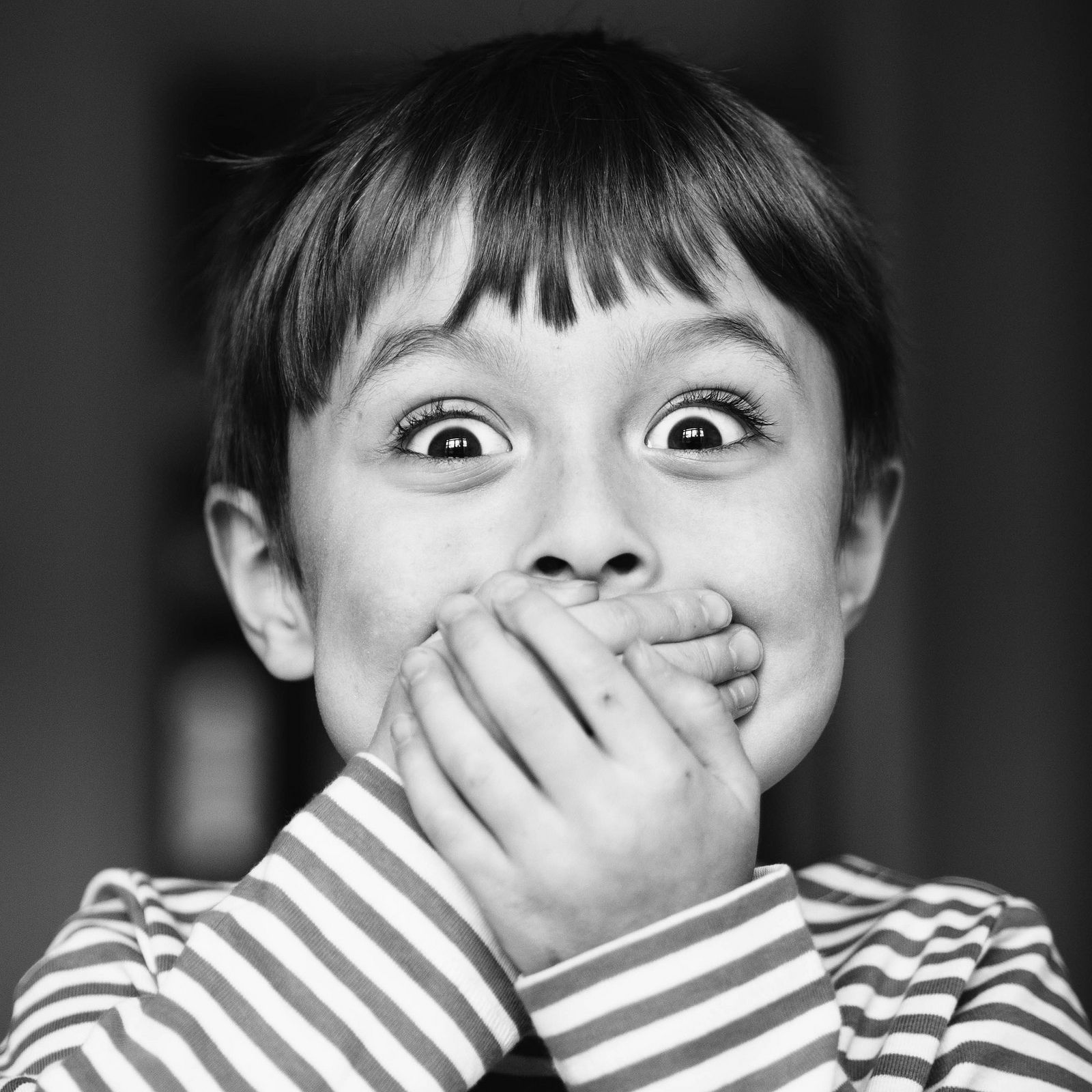 Black and white close-up of a child with a surprised expression, covering mouth with hands, wearing striped shirt.
