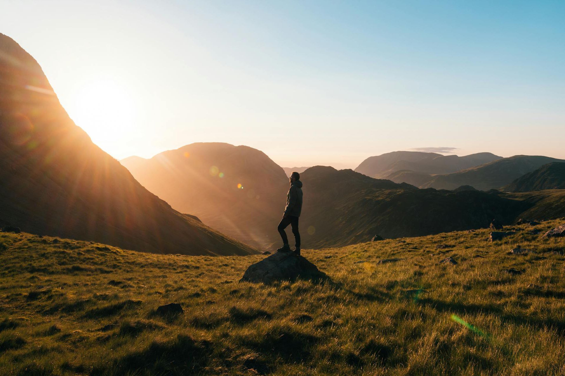Silhouette of a person standing on a grassy hill at sunrise, with mountains and warm light in the background