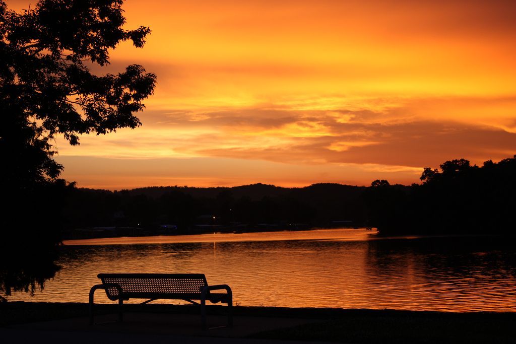 Sunset over a lake with a bench in silhouette. Orange sky reflects on the water.