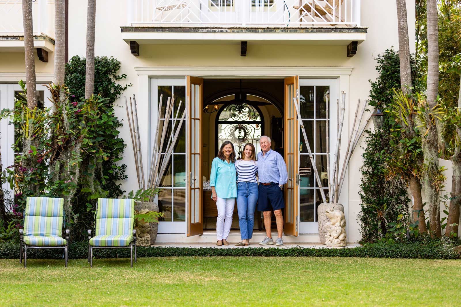 Three people stand in a doorway of a white house. Two chairs sit on the grass.