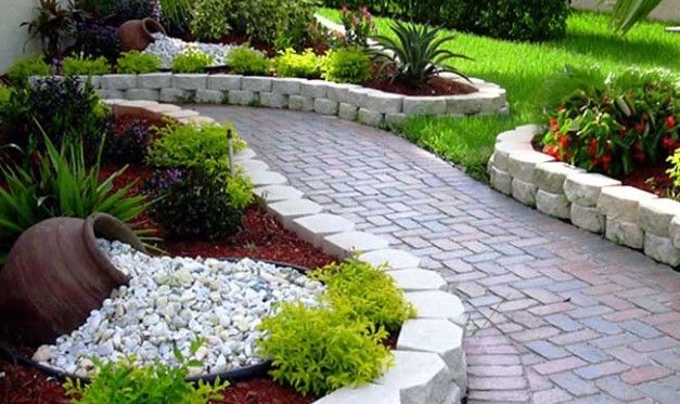 Curving brick pathway through a landscaped garden with raised beds, overflowing pots, and lush greenery.