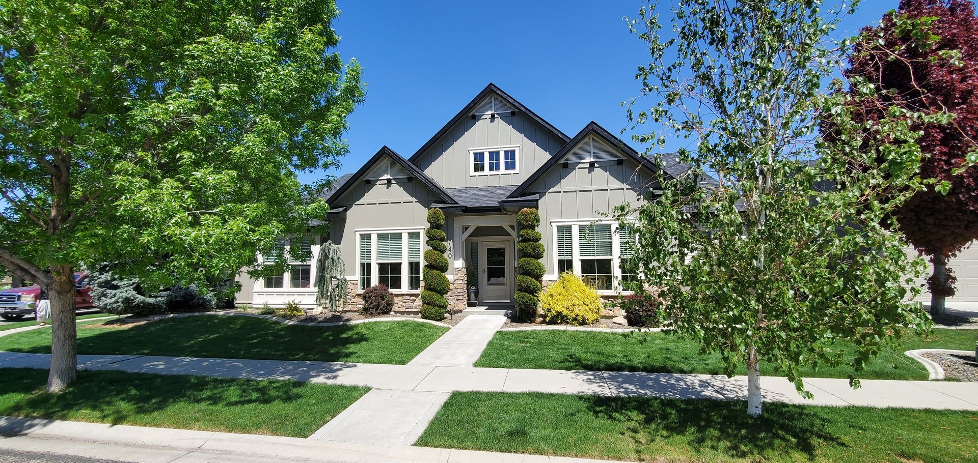 A light gray house with black trim has a walkway leading to the front door. Lush green grass, a blue sky, and trees surround the home.