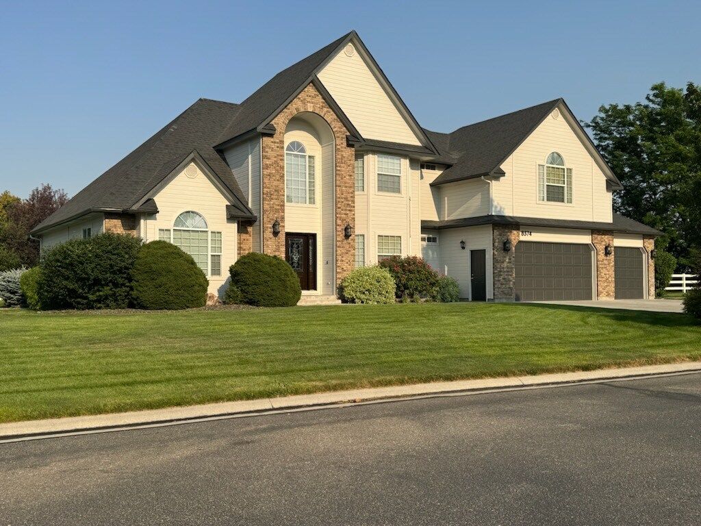 A large, two-story beige house with a dark roof, arched windows, and a well-manicured lawn. The house has a black front door and two garage doors.