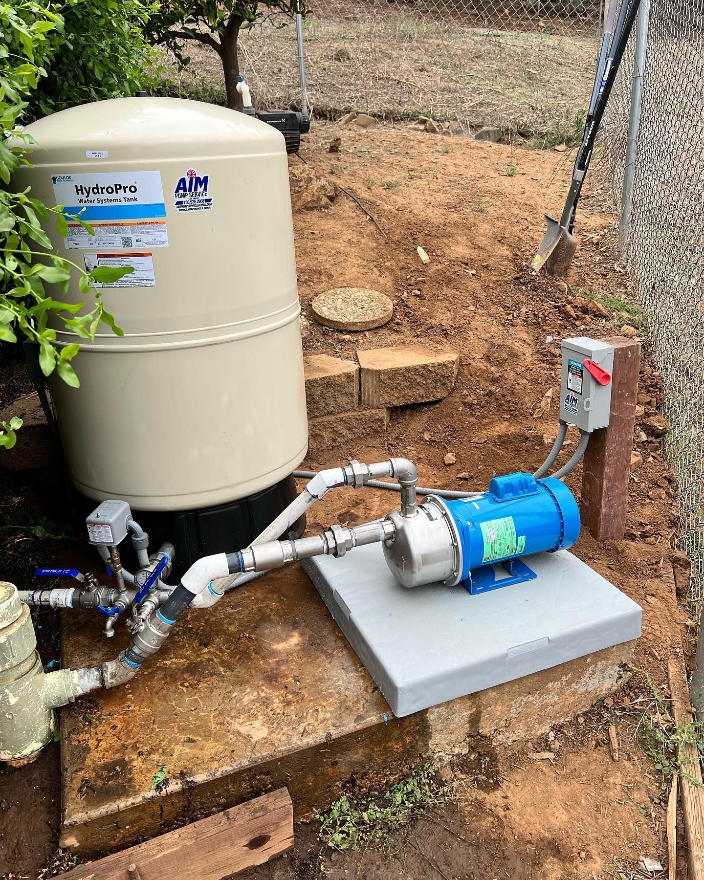 A light tan well pressure tank connected to a blue electric water pump on a concrete pad in an outdoor dirt area.