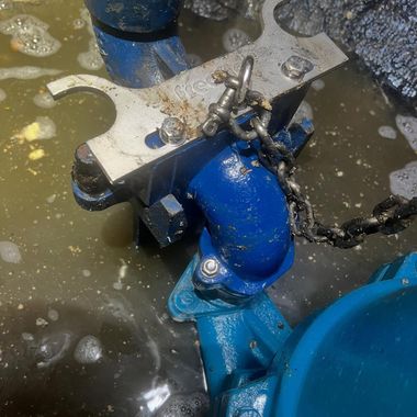 A blue industrial pump component partially submerged in murky water with a metal bracket and chain attached to it.