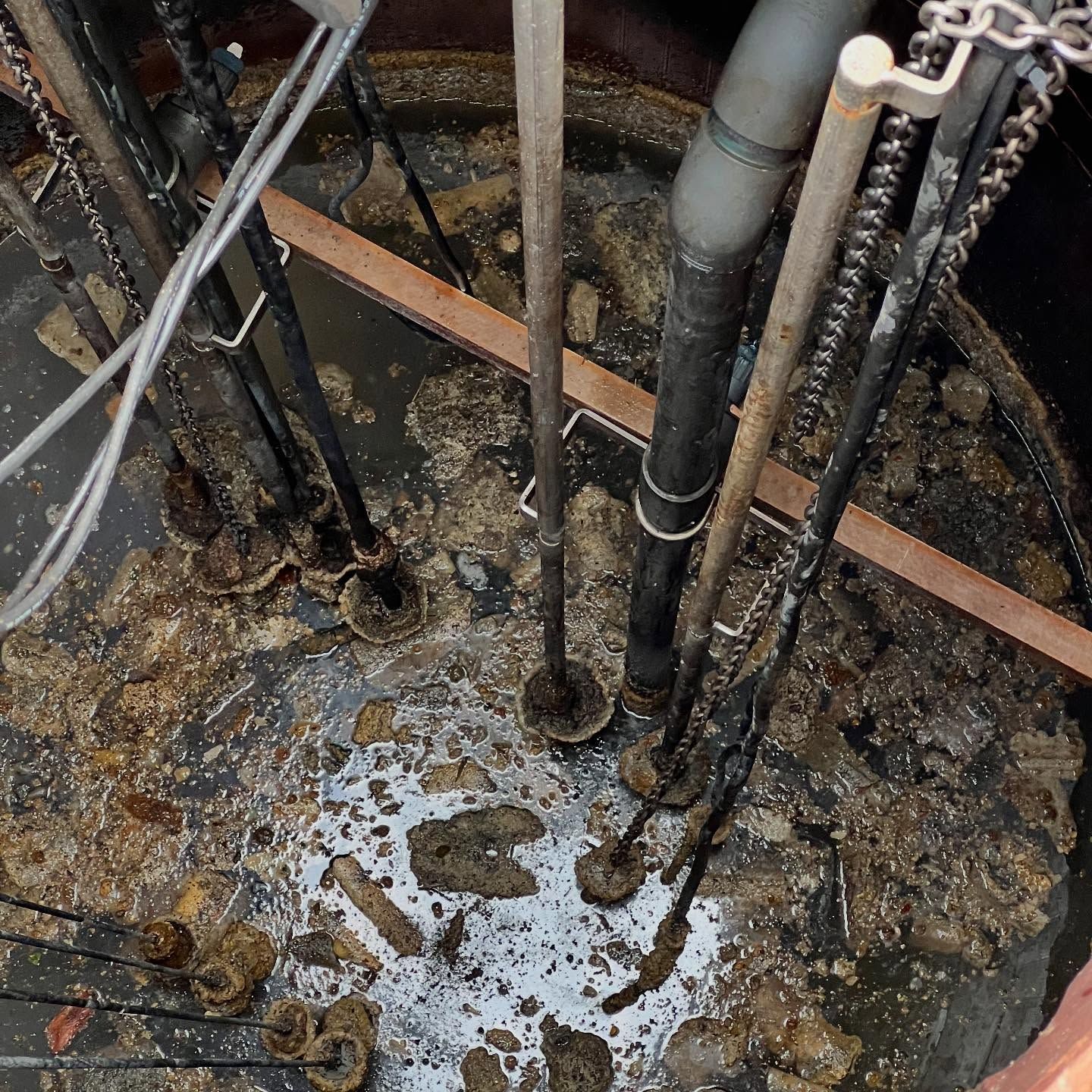 Top-down view of a dark, cluttered sewage pump station basin with suspended pipes, cables, and accumulated debris.