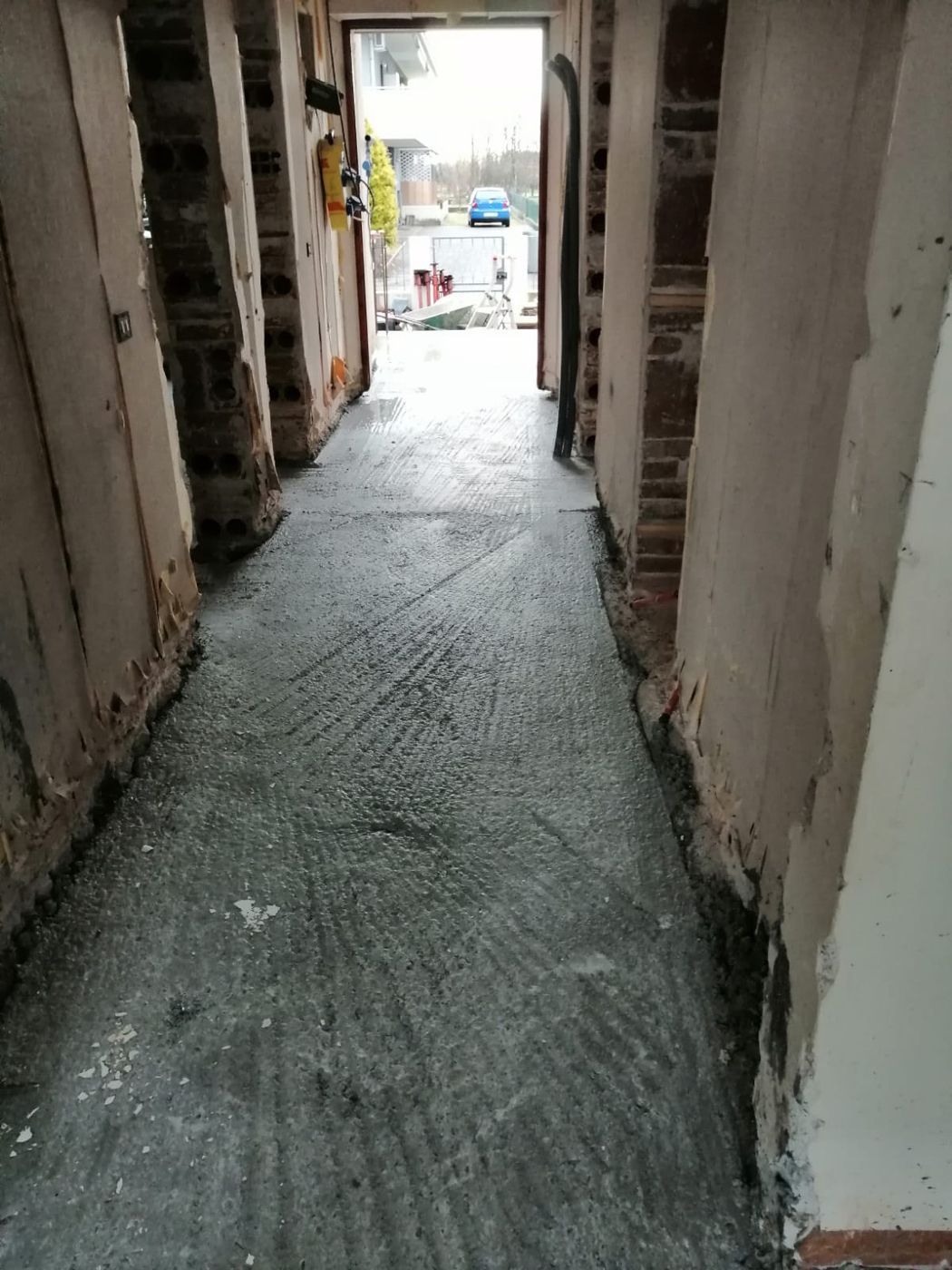 Un corridoio in fase di ristrutturazione con pareti in mattoni a vista, cavi elettrici scoperti e un pavimento in cemento appena posato.
