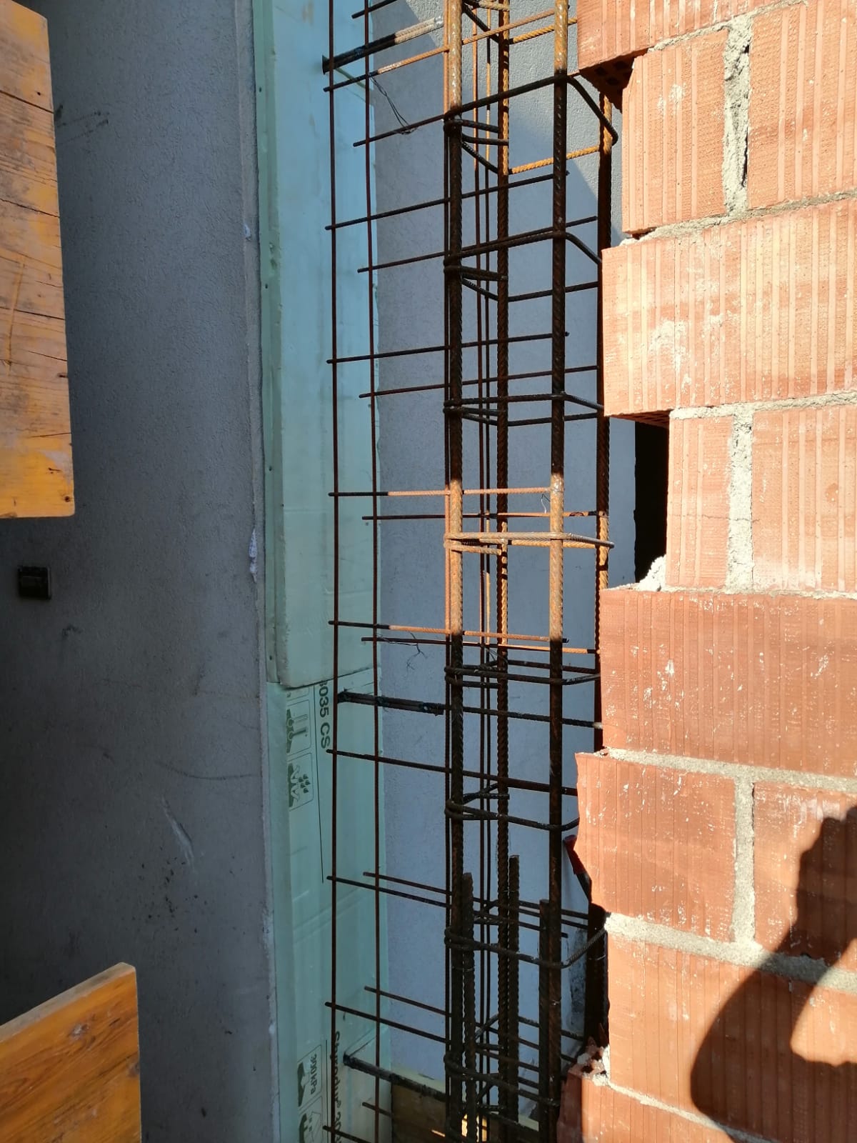In un cantiere edile, una gabbia di barre d'armatura si erge verticalmente tra una parete grigia e un muro di mattoni.