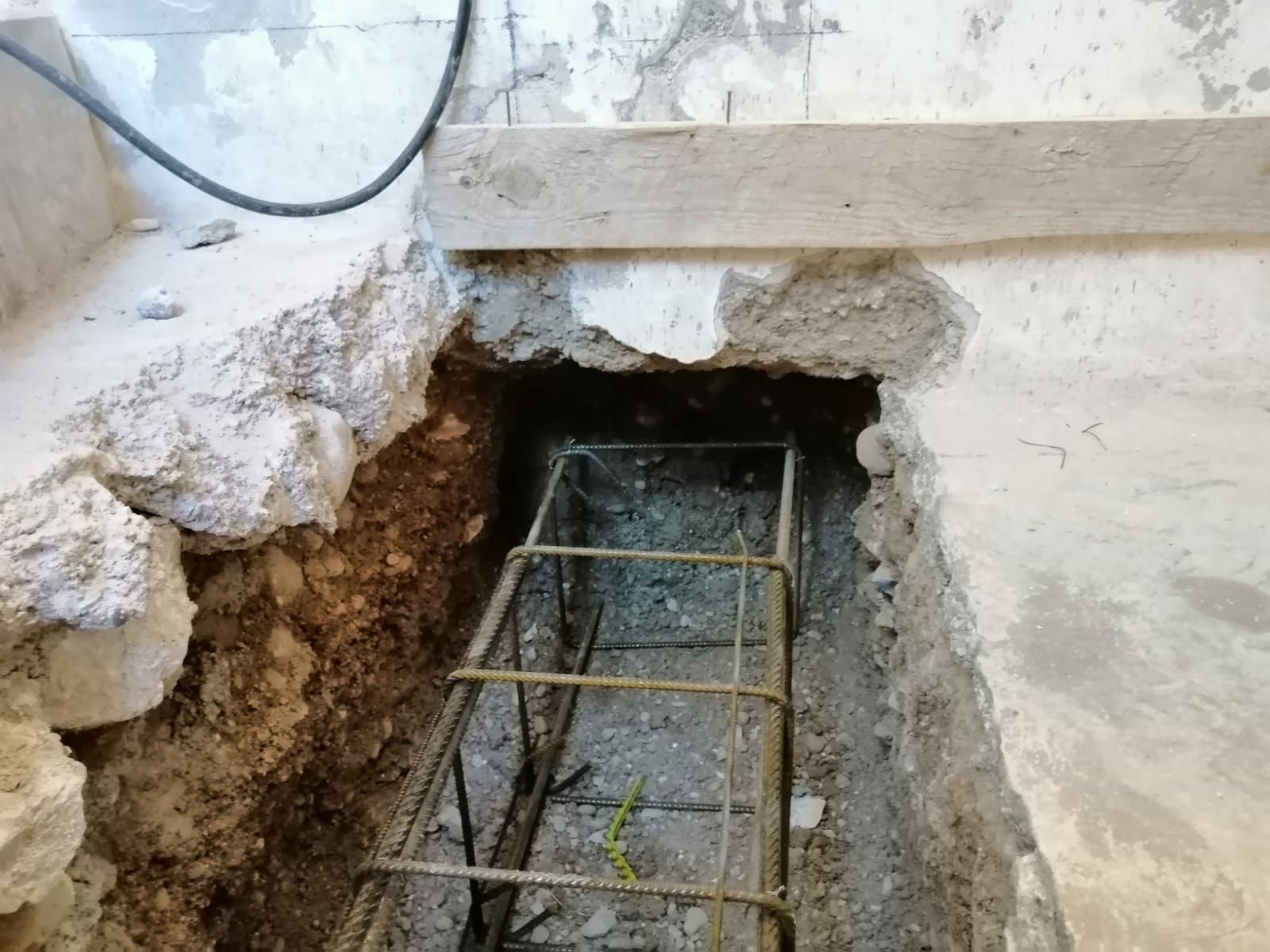 Una gabbia metallica per barre d'armatura è posizionata in una trincea scavata in un pavimento di cemento, predisposta per i lavori di consolidamento strutturale.