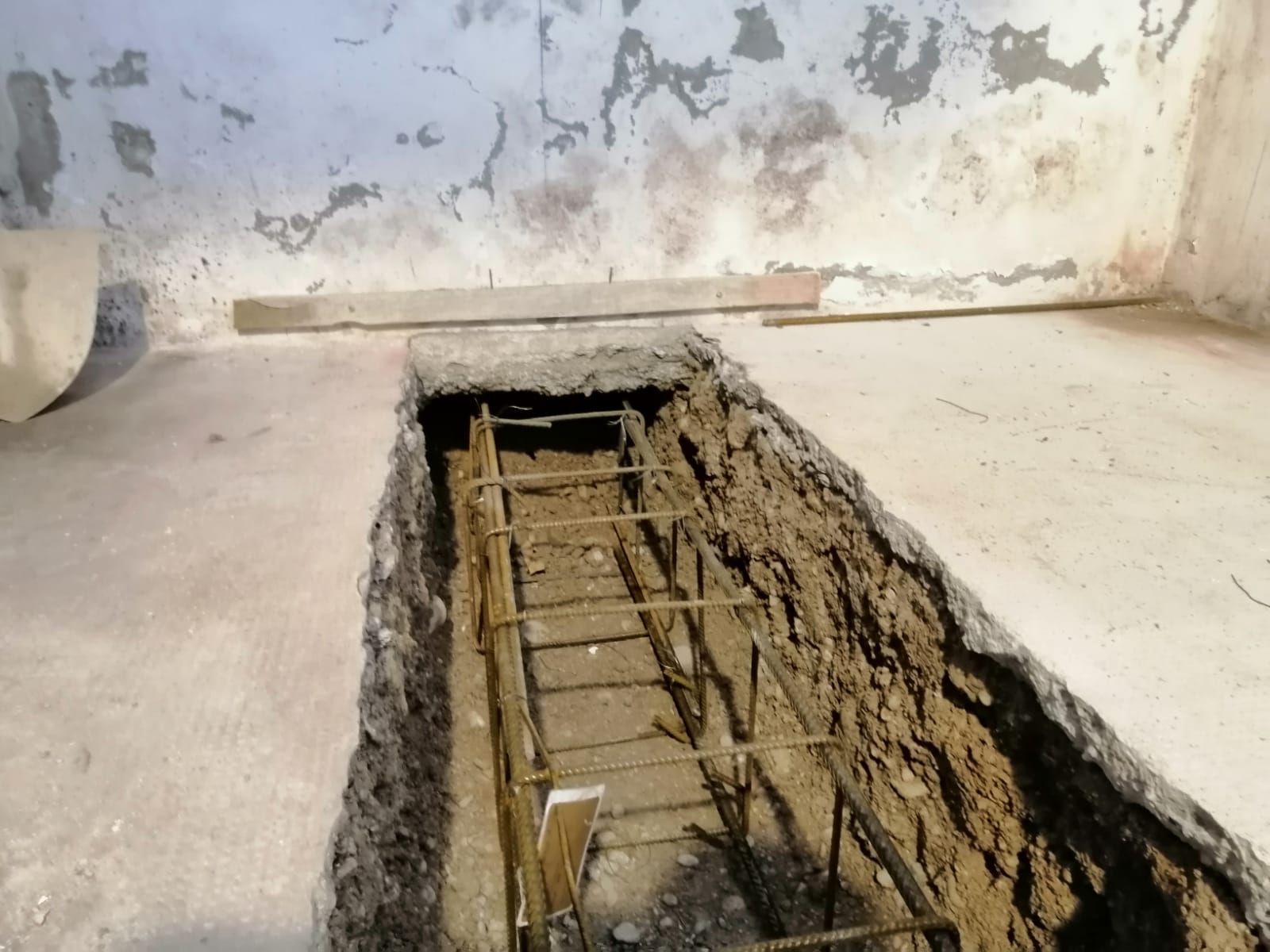 Una stretta trincea scavata in un pavimento di cemento rivela una gabbia di rinforzo in tondini metallici, probabilmente destinata a una trave di fondazione strutturale.