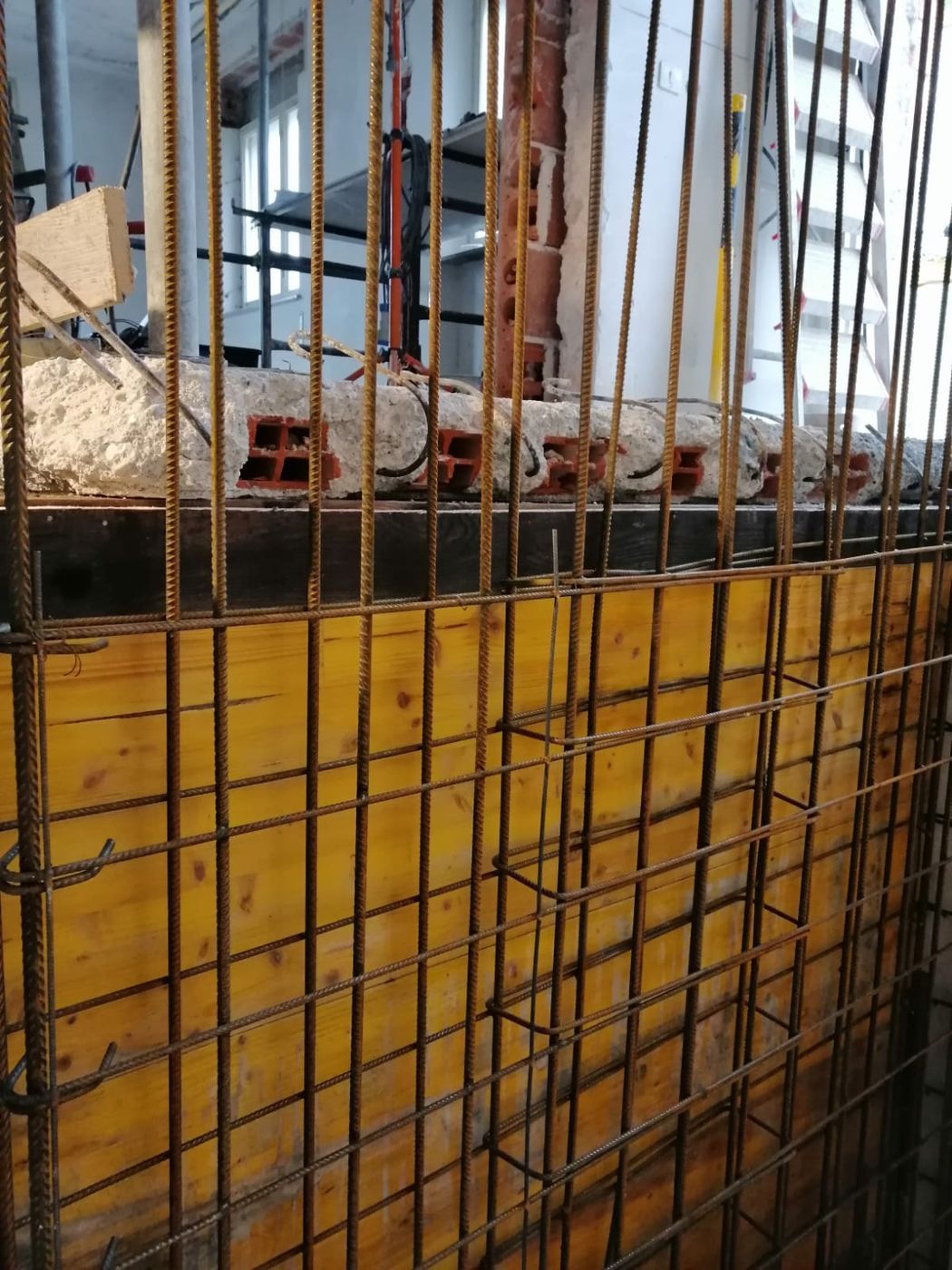 Un cantiere edile mostra un pannello di casseratura in legno giallo con una gabbia verticale di barre d'acciaio contro un soffitto rivestito di mattoni.