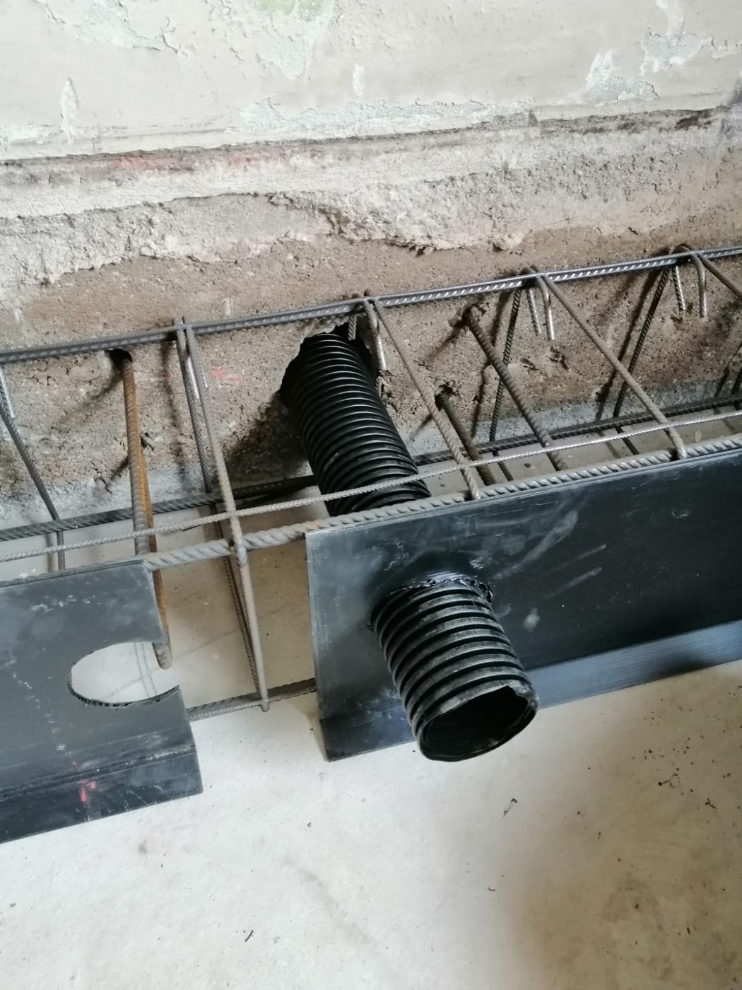 Un segmento di tubo corrugato nero inserito attraverso una gabbia di barre d'acciaio contro una parete di cemento grezzo.