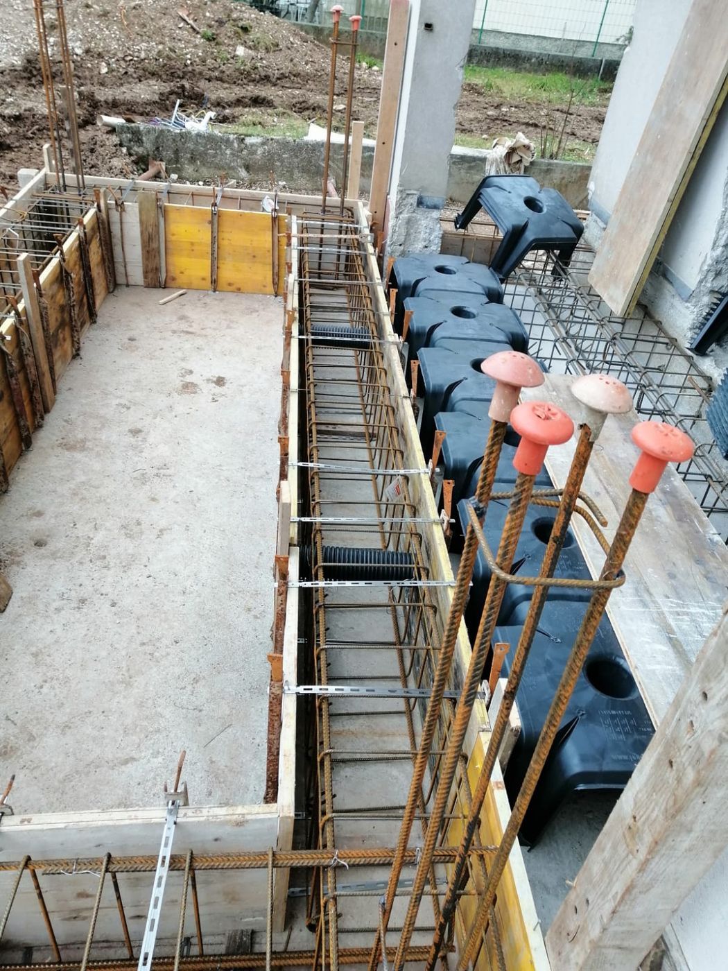 Cantiere edile che mostra una cassaforma in legno, una gabbia di barre d'acciaio per una trave e casseforme modulari in plastica nera.
