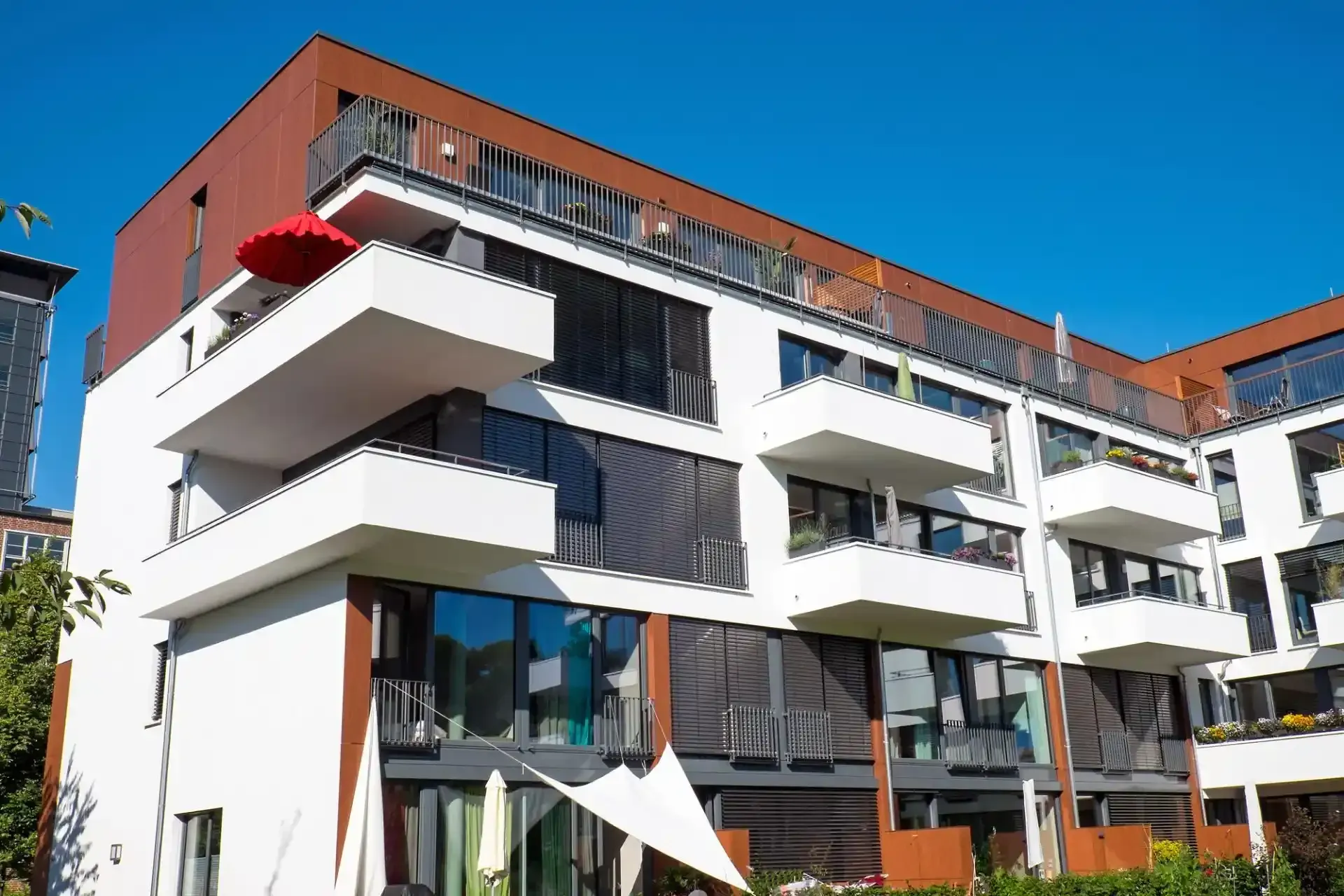 Ein modernes, mehrstöckiges Wohnhaus mit weißen Balkonen, großen Fenstern und einem braunen obersten Stockwerk unter einem klaren blauen Himmel.