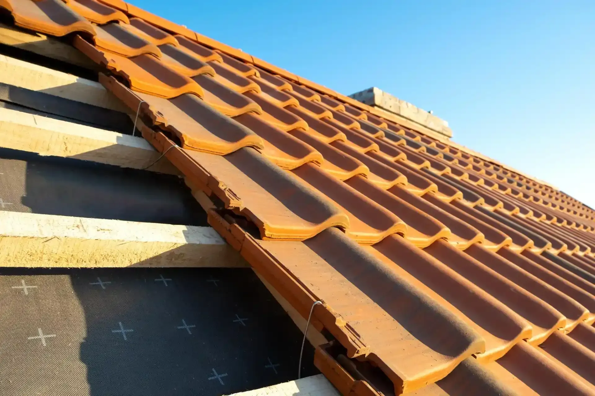 Posa di tegole in terracotta su una struttura di legno, sopra una membrana nera impermeabile, sullo sfondo di un cielo azzurro.