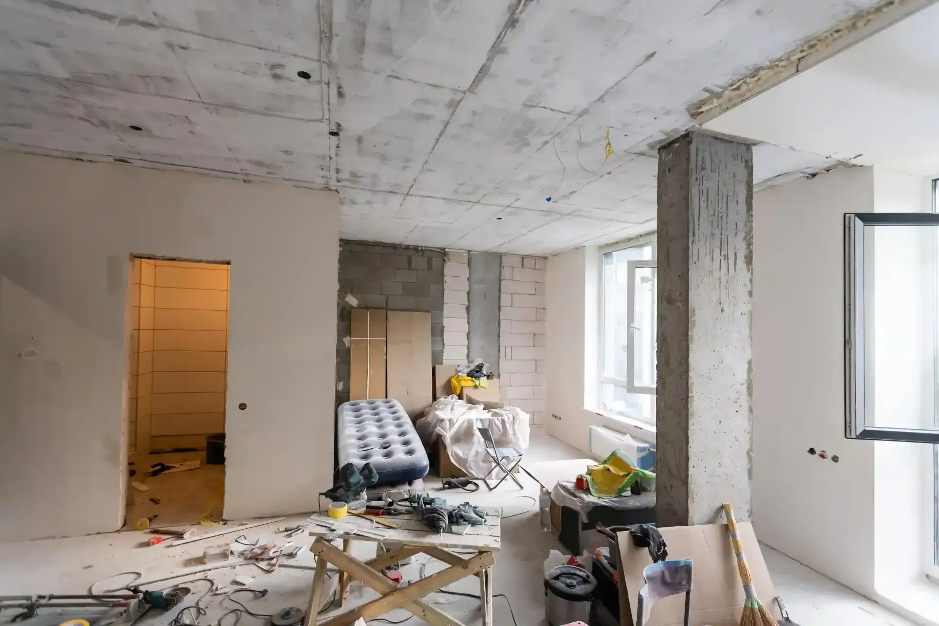 Innenansicht eines im Bau befindlichen Raumes mit nackter Betondecke, unfertigen Wänden, Werkzeugen und herumliegenden Materialien.