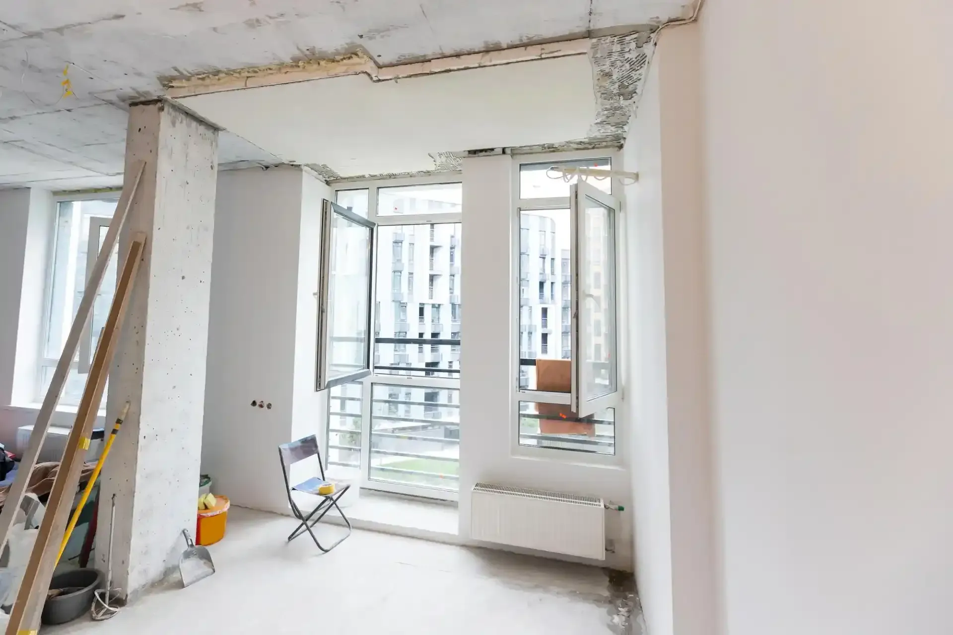 Una stanza non finita, in fase di ristrutturazione, con pareti bianche, soffitto in cemento, una sedia pieghevole e finestre che si affacciano sulla città.
