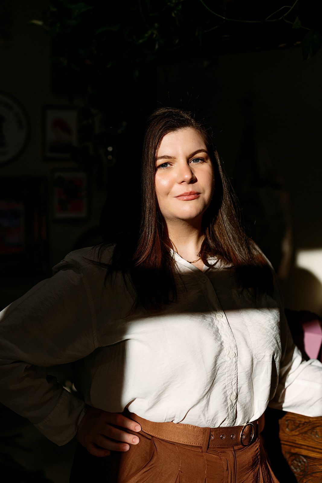 Woman standing indoors with hands on hips, wearing a white blouse and brown pants, lit by sunlight